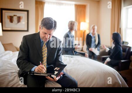 Businessman sits on the edge of a bed in a hotel room during a business trip and writes in a folder while his three female colleagues work together in the background. Stock Photo