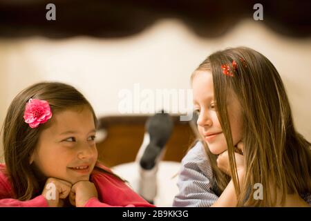 Two young girls smile at each other as they lie front down on a bed together and prop themselves up on their elbows to pose for a portrait. Stock Photo