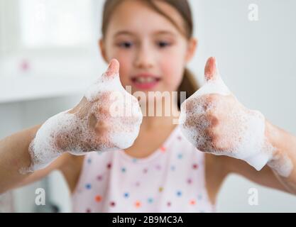 Little girl washing hands with water and soap in bathroom. Kid showing thumbs up. Hands hygiene and virus infections prevention. Stock Photo