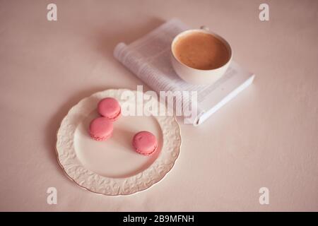 Pink macaroon cookies on white plate with open book and cup of coffee in bed closeup. Good morning. Breakfast time. Selective focus. Stock Photo