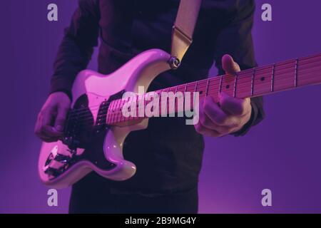 Young and joyful caucasian musician playing guitar on gradient purple studio background in neon light. Concept of music, hobby, festival. Colorful portrait of modern artist. Attented and inspired. Stock Photo