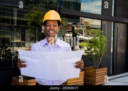 Portrait of professional architect in helmet looking at blue prints outside modern building. Engineer and architect concept. Stock Photo