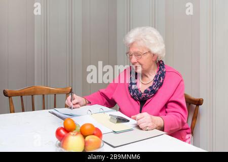 Old woman sits at the table over a file folder and uses calculator. Numbers and notes in the background Stock Photo
