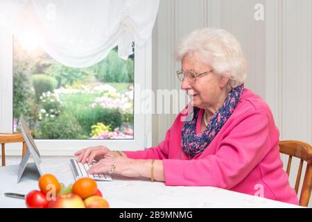 80 year old woman sitting alone at home. She belongs to the risk group for Covid 19. Senior woman writes on the tablet PC against isolation with her f Stock Photo