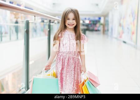 Kid holding shopping bags. Little girl in pink dress looking through shop window. A smiling laughing girl in a pink dress with multi-colored bags in her hands is walking around the mall, looking at the shop windows. Stock Photo