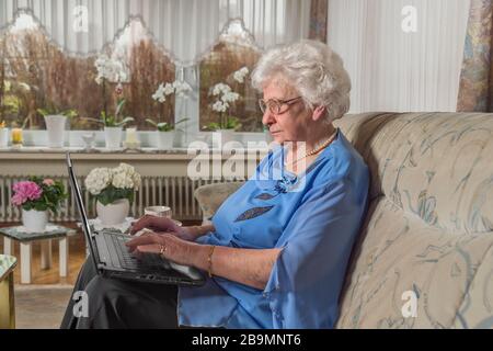 80 year old woman sitting alone at home. She belongs to the risk group for Covid 19. Senior woman writes on the laptop against isolation with her fami Stock Photo