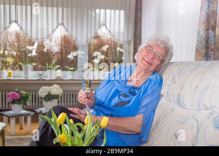 80 year old woman sitting alone in the apartment. She belongs to the risk group for Covid 19. She drinks a glass of sparkling wine and distracts herse Stock Photo