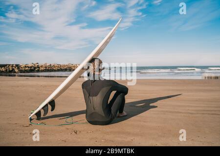Young surfer sitting on sandy beach and next to his surfboard. Sport and water sport concept. Stock Photo