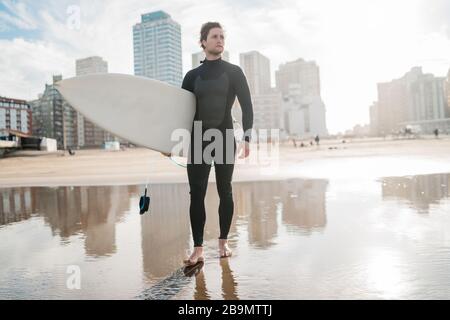 Young surfer standing in the ocean with his surfboard in a black surfing suit. Sport and water sport concept. Stock Photo