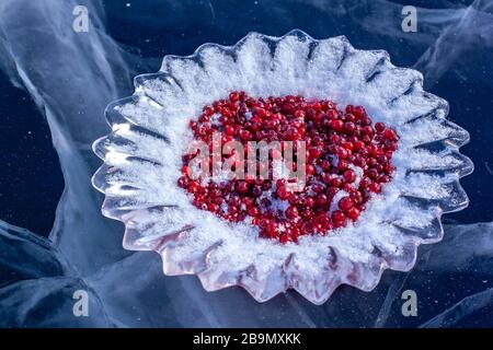 An ice plate with cranberries sprinkled with snow stands on the ice of Lake Baikal. Beautiful cracks in the ice. Red berries on ice-cut dishes. Russia Stock Photo