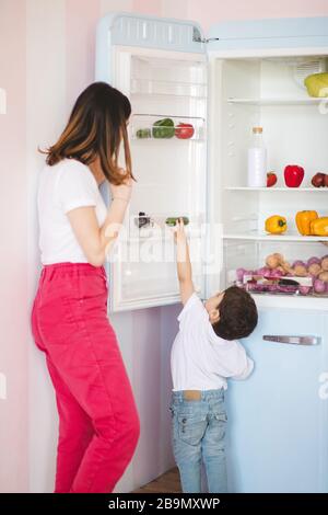 Little cute boy trying to pick something from fridge while his mother opening the fridge door. Stock Photo