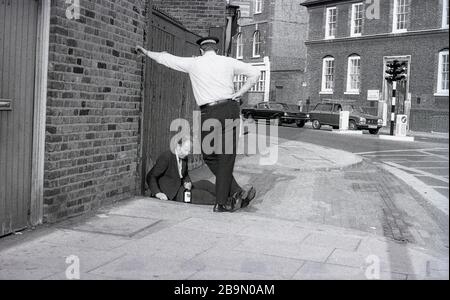 1973, historical, a British police constable or 'bobbie' standing talking to a man sitting on the pavement  slumped with a bottle of drink in his hand, London, England, UK. In this era and previous decades, the local 'Bobbie' would walk or patrol his territory or beat and know the surroundings and local people. This traditional form of policing, which was based on a close relationship with the community, changed in later decades, as the police became a force that operated out of cars and not on foot. Stock Photo