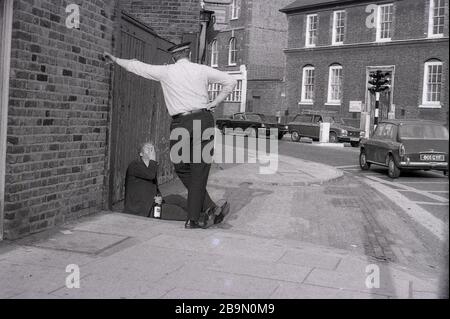 1973, historical, a British police constable or 'bobbie' standing talking to a man sitting on the pavement  slumped with a bottle of drink in his hand, London, England, UK. In this era and previous decades, the local 'Bobbie' would walk or patrol his territory or beat and know the surroundings and local people. This traditional form of policing, which was based on a close relationship with the community, changed in later decades, as the police became a force that operated out of cars and not on foot. Stock Photo