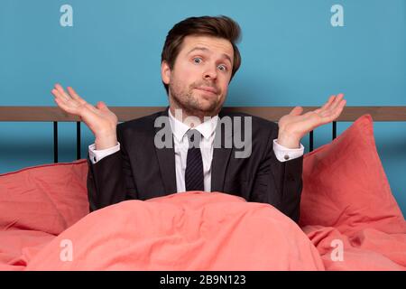 Doubtful young man sitting in bed and gesturing with hands with doubt being unsure in future. isolated on bluebackground Stock Photo