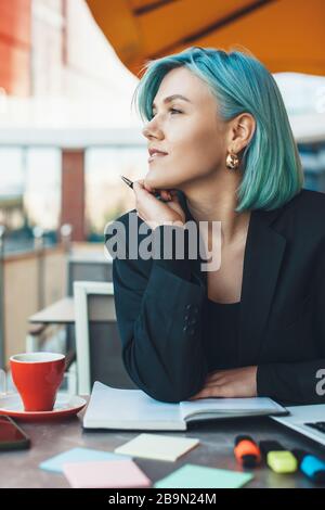 Pensive caucasian blue haired lady looking away while sitting in a coffee shop and making some notes Stock Photo