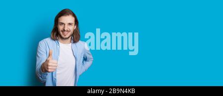 Long haired caucasian man with beard is gesturing the like sign on a blue wall with copy space Stock Photo