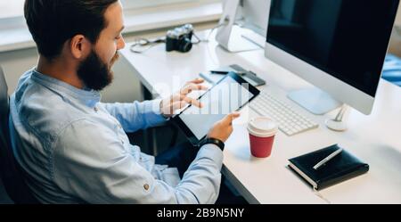 Upper view portrait of a bearded caucasian man using a tablet with blank space while drinking a coffee in his office Stock Photo