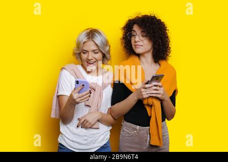 Blonde caucasian woman holding a phone and posing near her curly haired friend on a yellow wall Stock Photo