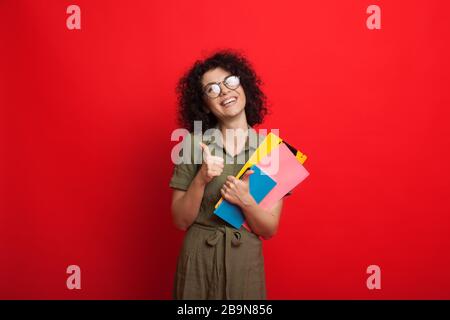 Curly haired caucasian student is holding some books and gesturing the like sign while posing on a red background Stock Photo