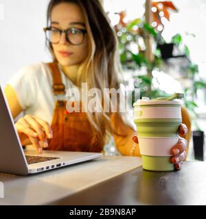 Young female gardener in glasses using laptop, communicates on internet with customer in home garden/greenhouse, hold reusable coffee/tea mug.Cozy off Stock Photo