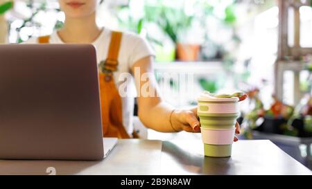 Female freelancer holding reusable coffee/tea mug, using laptop, communicates on internet in home garden/greenhouse. Selective focus. Cozy office work Stock Photo