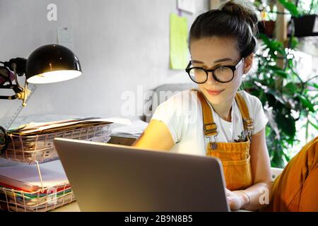 Young female gardener in glasses using laptop, communicates on internet with customer, works from home during quarantine due to coronavirus. Cozy offi Stock Photo