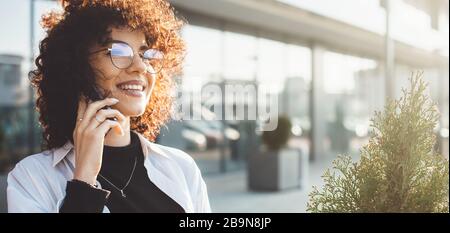 Curly haired businessperson having a phone discussion while wearing eyeglasses and posing outside Stock Photo