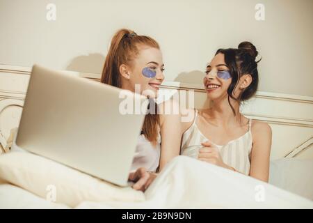 Two caucasian sisters wearing some anti aging masks while lying in the bed with a computer Stock Photo