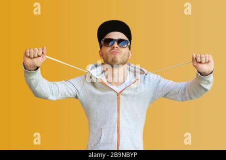 young hipster in sunglasses wearing a cap against a yellow wall in the street. A guy in a white T-shirt, sweater and beard is grimacing at the camera. Stock Photo