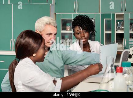 Female African medical students or young graduates discuss with Caucasian senor male group leader in research or medical laboratory. Looking for treat Stock Photo