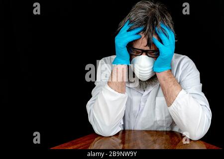 A man wearing a mask, blue latex gloves, and white lab or doctor coat holds his head in his hands. He looks tired and frustrated. Black background. Stock Photo