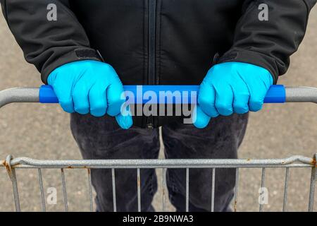 A person wearing blue latex gloves pushes a shopping cart in a parking lot. Closeup view focused on the hands. Stock Photo