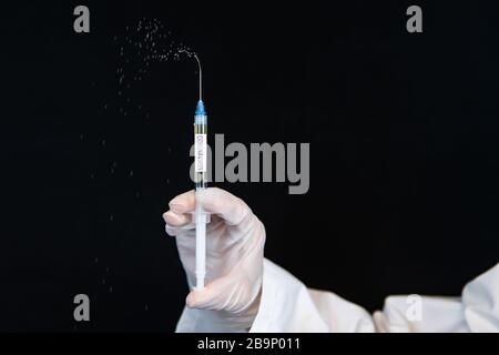 Selective focus closeup, syringe labeled coronavirus, squirting drops of liquid being held by a gloved hand vertically over a black background  Stock Photo