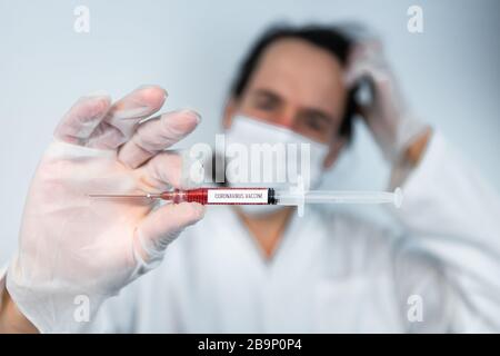 Closeup of syringe filled with red liquid with label coronavirus vaccine held horizontally center frame by confused out of focus medical professional  Stock Photo