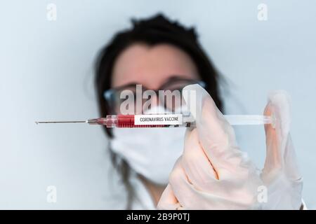 Selective focus shot of syringe held by gloved hand horizontally in center frame filled with red fluid. Background out of focus nurse wearing glasses Stock Photo