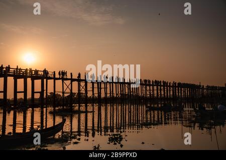 The U-Bein bridge at sunset, Mandalay, Myanmar. People silhouetted on long wooden bridge against colourful sky and water background Stock Photo