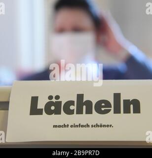 24 March 2020, Saxony, Leipzig: In the office of the association 'Alter, Leben und Gesundheit' in one of the longest blocks of flats in Germany, the 'Lange Lene', a female employee with a mouthguard sits at a desk with the sign 'Smiling simply makes you more beautiful'. With face masks and protective gloves, more than 200 elderly people in the 335-meter-long prefabricated building of the Leipziger Wohnungsbaugesellschaft (LWB) are cared for by the association's employees, also as a small family substitute. In order to ensure that lunch is safe because the cafeteria is now closed, some of them Stock Photo
