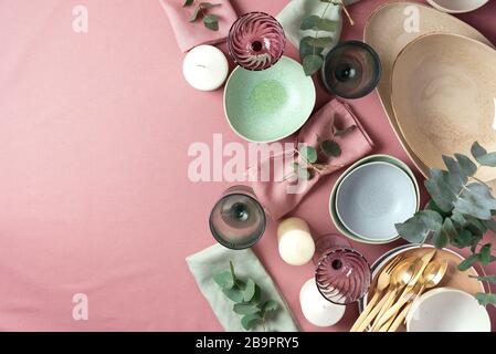 Table served for festive spring dinner. Top view. Copy space. Empty serving plates, bowls, candles and eucalyptus flowers on pink shabby background Stock Photo