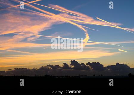 Colorful contrails high up in the sky are illuminated by the light of the setting sun. Closer to the surface of the earth, there are dark clouds. Stock Photo