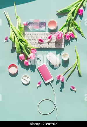 Female home office. Pastel blue desktop with PC keyboard , smartphone with blank screen and wire , cosmetics and pink tulips flowers in sunlight. Top Stock Photo