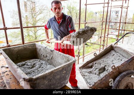 Construction worker is using shovel to unloading cement, mortar, from wheelbarrow on construction site. Stock Photo
