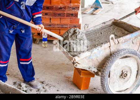 Construction worker is using shovel to unloading cement, mortar, from wheelbarrow on construction site. Stock Photo