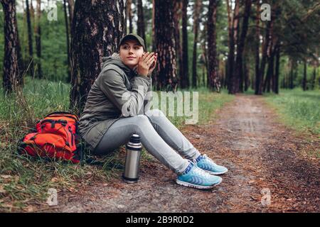 Woman tourist drinks hot tea from thermos cup relaxing in spring forest with backpack. Camping, traveling and sport Stock Photo