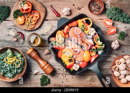 Salmon steak with vegetables on iron grill pan, bowl with kale salad, nuts, organic vegetable and kitchen utensils over wooden background. Top view. F Stock Photo