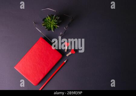 A red notebook stands on a black background, next to a green plant notebook, glasses, a red pencil and two heart-shaped pins Stock Photo