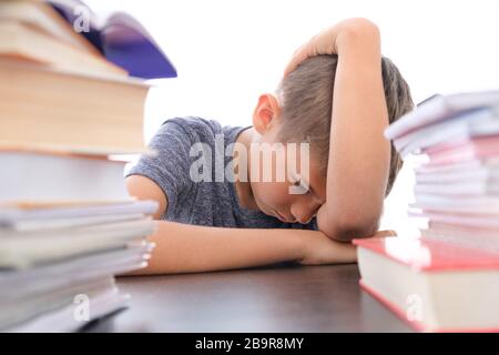 Tired disappointed schoolboy lowering his head sitting among pile of books, textbooks, school exercise books on his desk at home Stock Photo