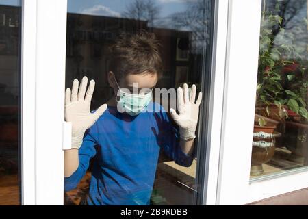 boy at home in quarantine with mask looks out of the closed window to protect himself from the pandemic of n-cov19 Coronavirus Stock Photo