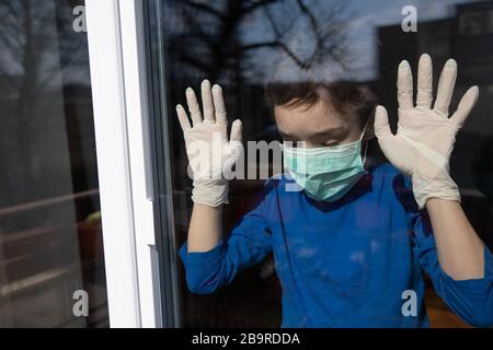 boy at home in quarantine with mask looks out of the closed window to protect himself from the pandemic of n-cov19 Coronavirus Stock Photo