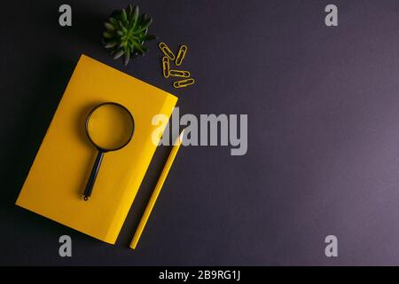 Yellow pencil, yellow notebook glasses, pins, clip paper and a magnifying glass on on black background Stock Photo