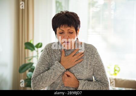 Sick man wearing scarf lying on couch at home under a blanket checking temperature. Living room with table full of medicine and pills. Stock Photo